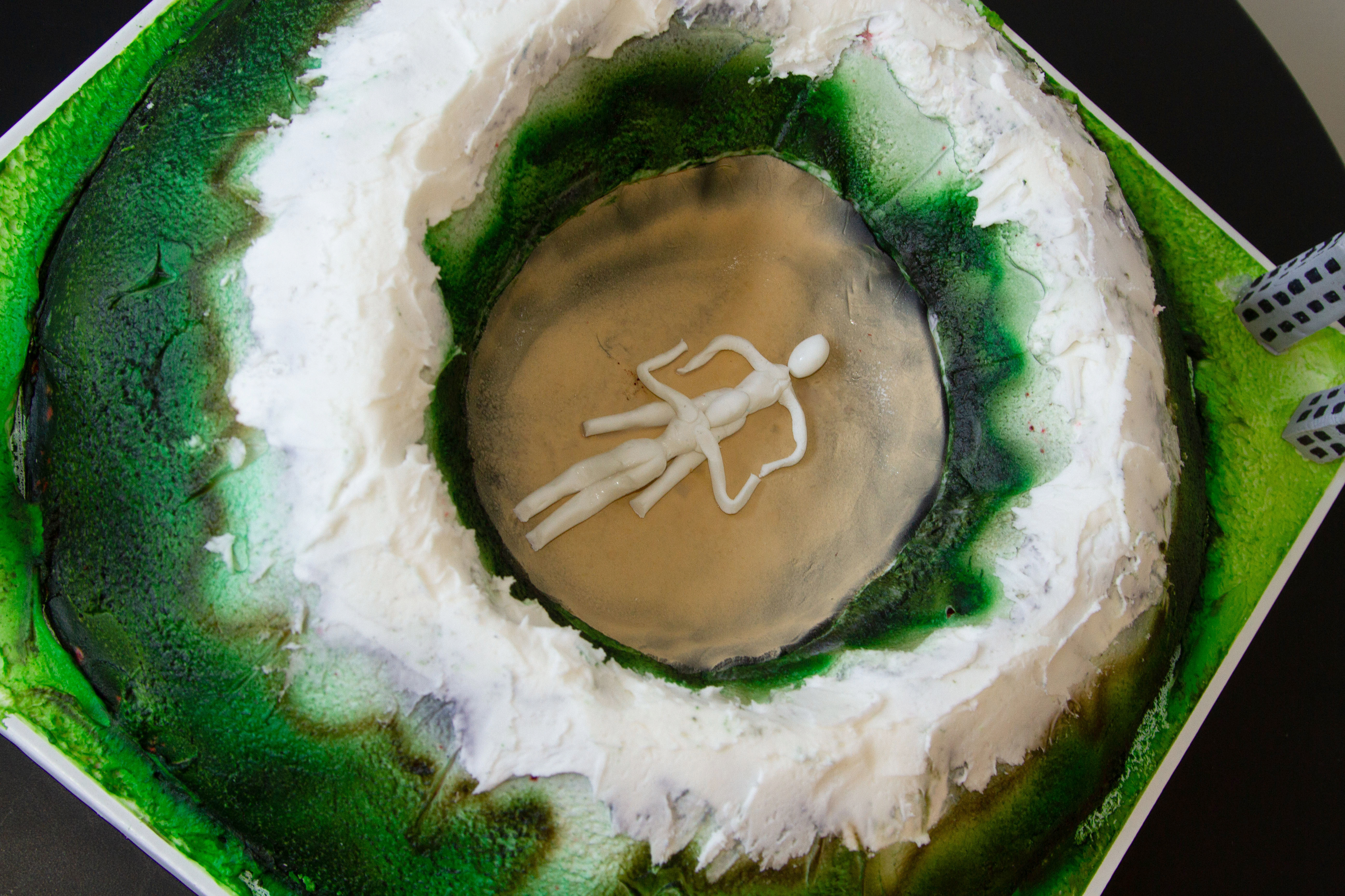 Detail ariel view of a cake shaped like a mountainous crater with two figures engaged in felatio in the center